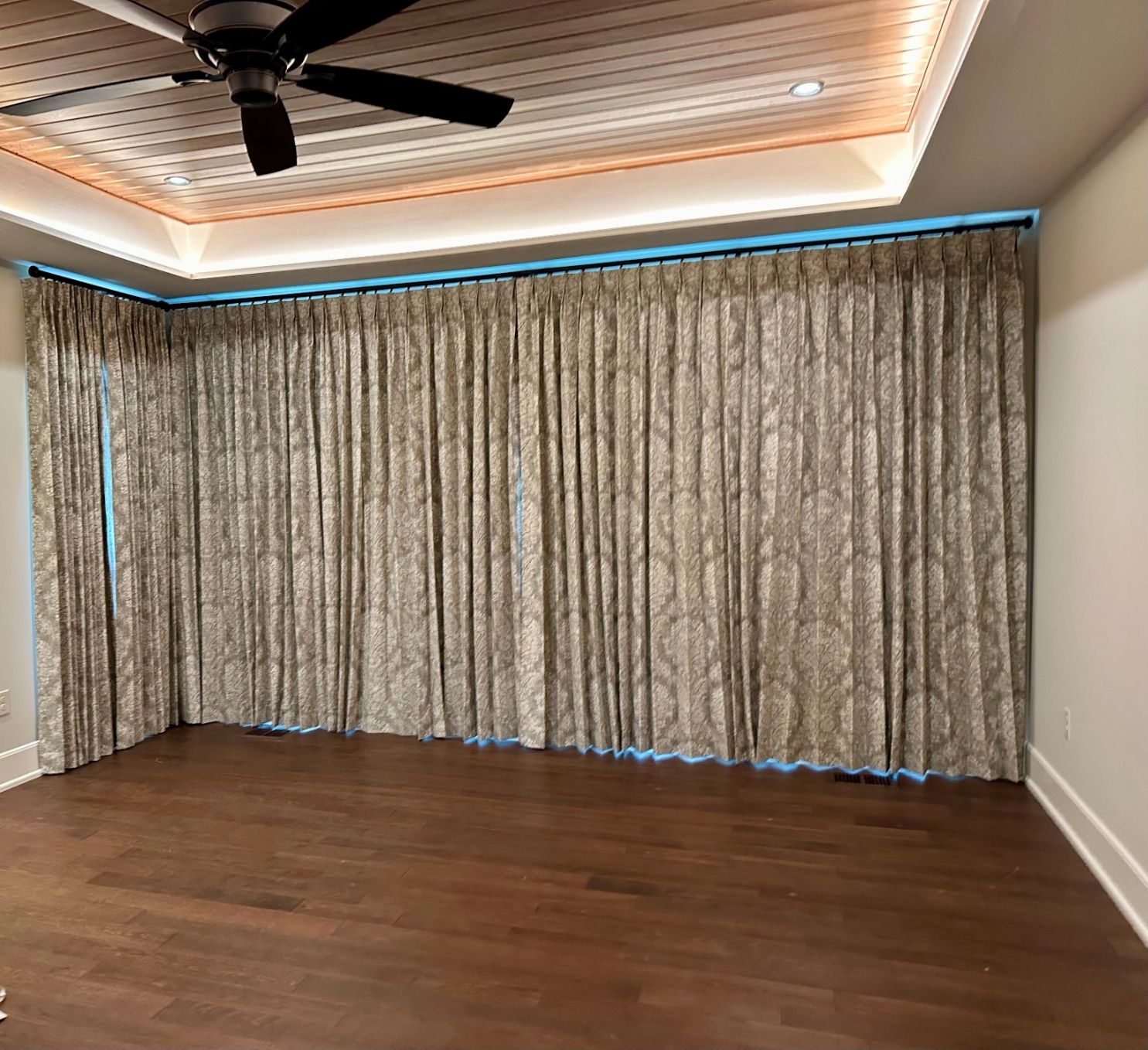 Empty room with wood floor, beige curtains, and a wood-paneled ceiling with a ceiling fan.