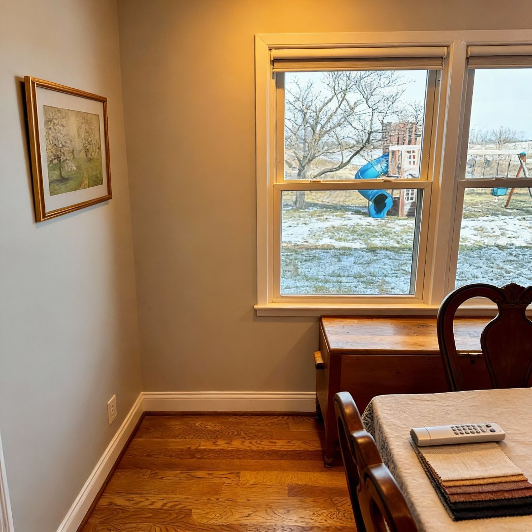 Room corner with wood floor, window, painting, table set with fabric swatches. Outside is a backyard.