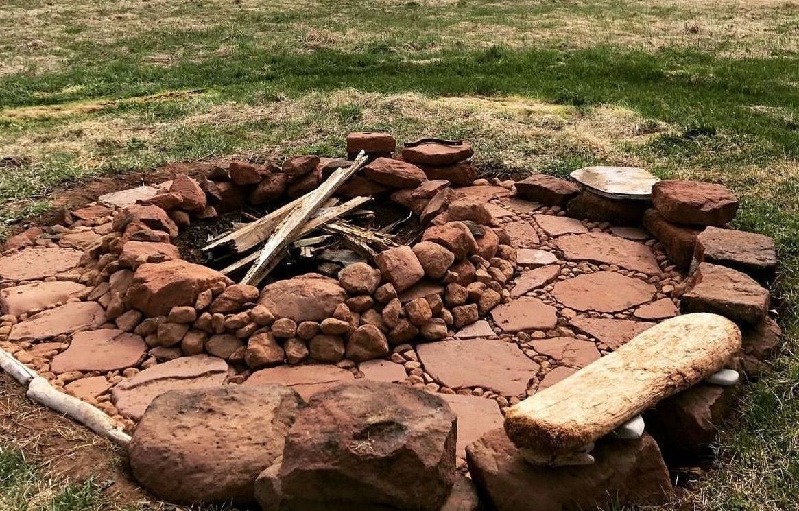 Fire pit with stacked brown stones, surrounded by red paving stones on grass. Wood inside.