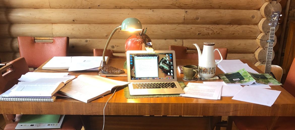 Desk with open laptop, documents, lamp, and coffee pot in front of a log cabin wall.