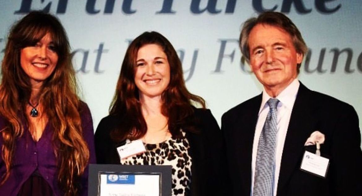Three people smiling, posed for a photo. A woman in purple, a woman in a black and white top, and a man in a suit.
