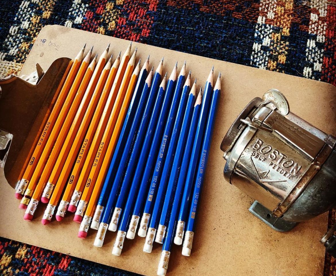 Pencils, orange and blue, next to a vintage Boston pencil sharpener on a clipboard.