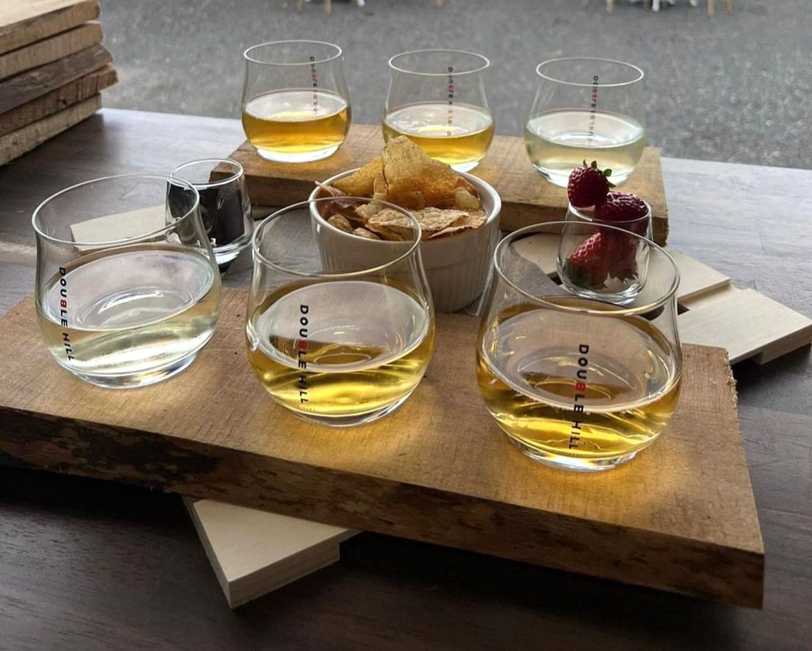 Flight of drinks on a wooden board with small snacks and garnishes.