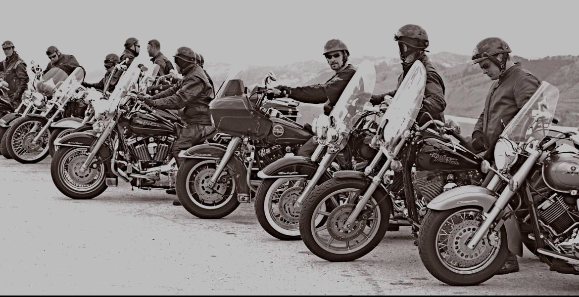 Motorcyclists in helmets lined up on bikes, with a mountain backdrop.