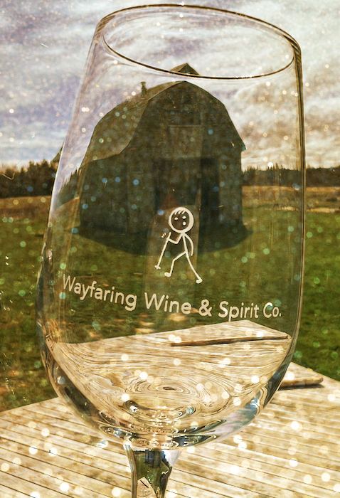 Wine glass with the logo of Wayfaring Wine & Spirit Co. in front of a rustic barn.