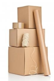 A stack of three cardboard boxes with a roll of brown packing paper, a roll of tape, and twine sitting on top.