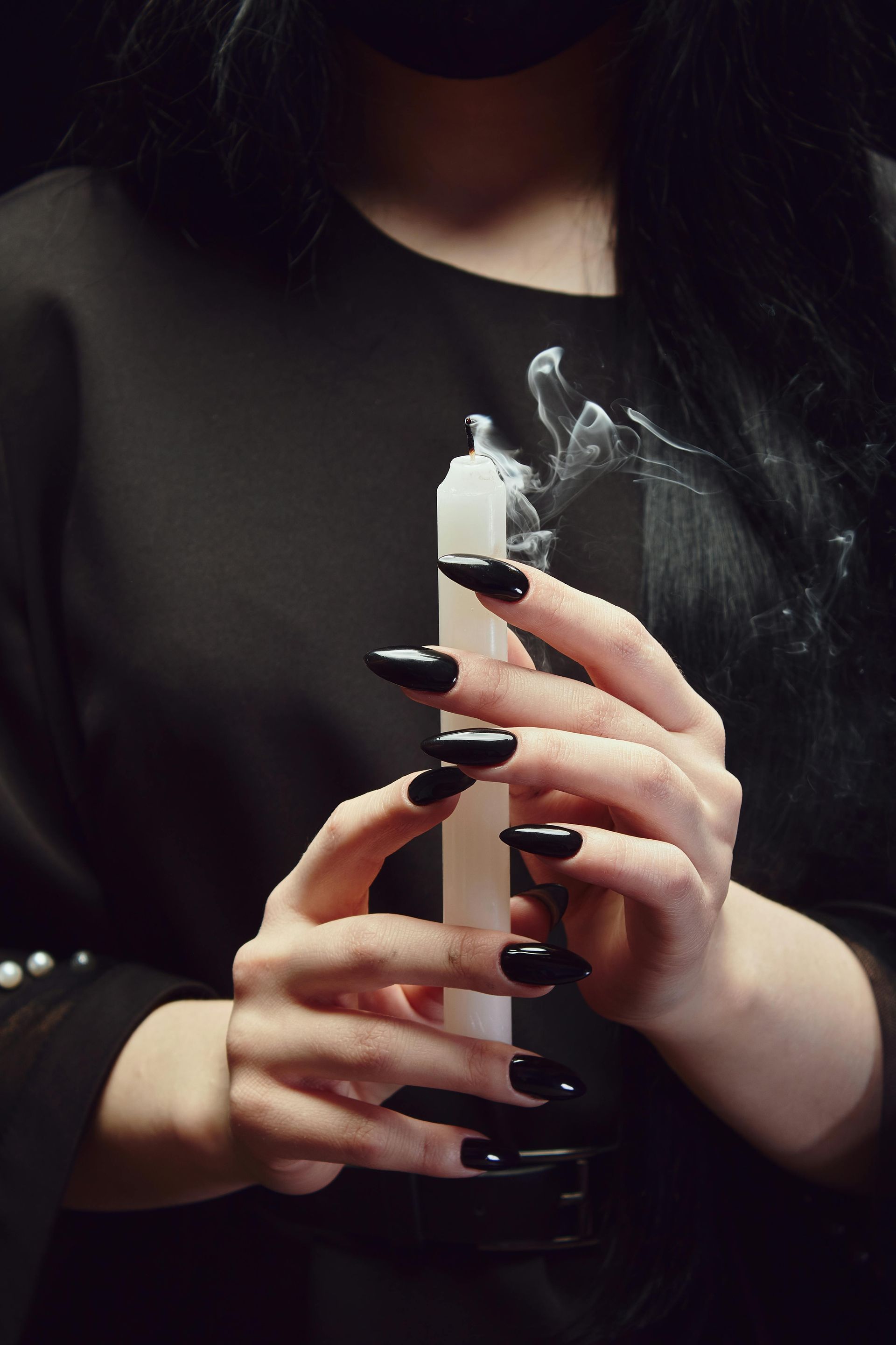 Vrouw met zwarte nagels die een rokende witte kaars vasthoudt tegen een zwarte achtergrond.