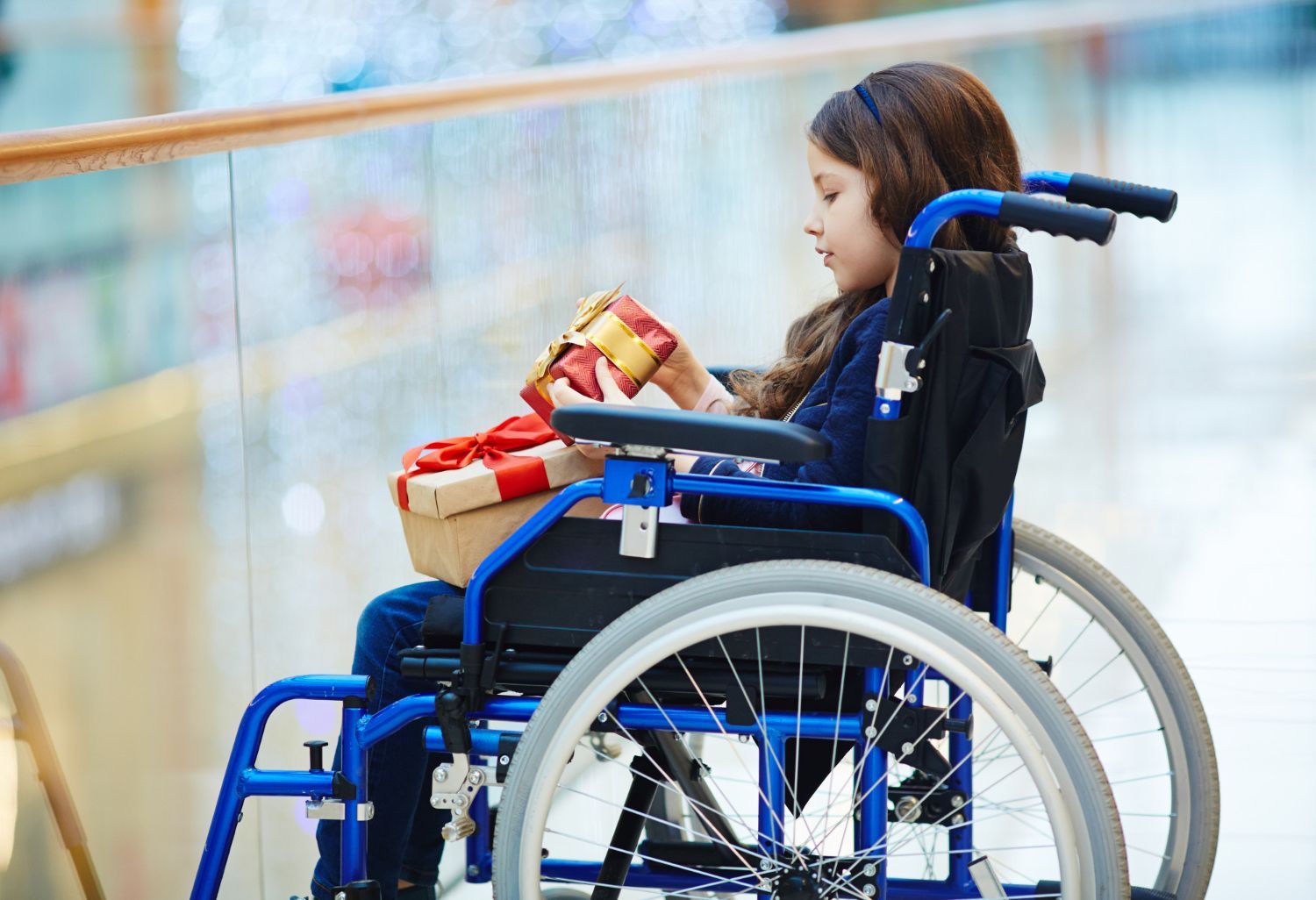 Child in a blue wheelchair holding a toy, outdoors near a railing and blurred background.