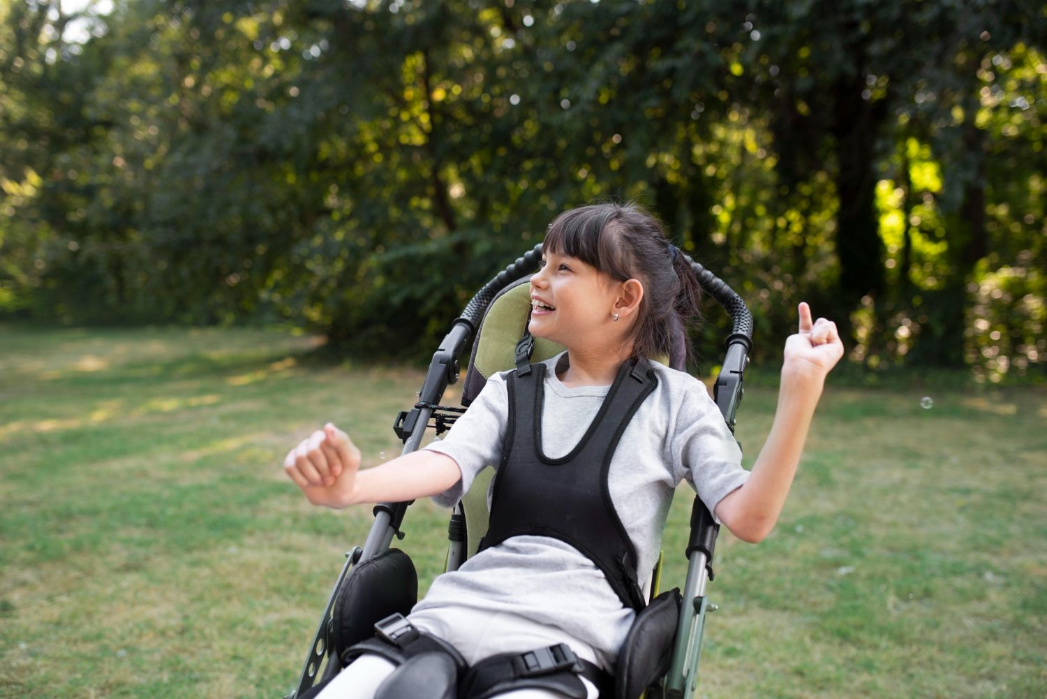 Child in a wheelchair smiling and raising both hands in a sunny park