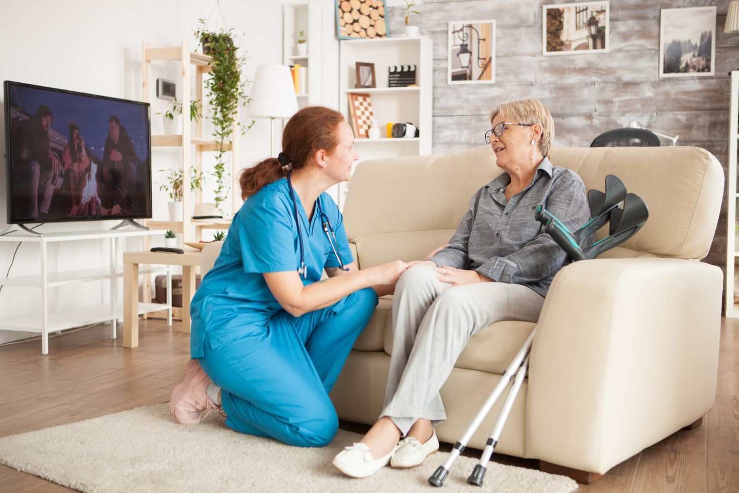 Caregiver in blue scrubs talks with elderly woman on a sofa in a bright living room, with crutches nearby.