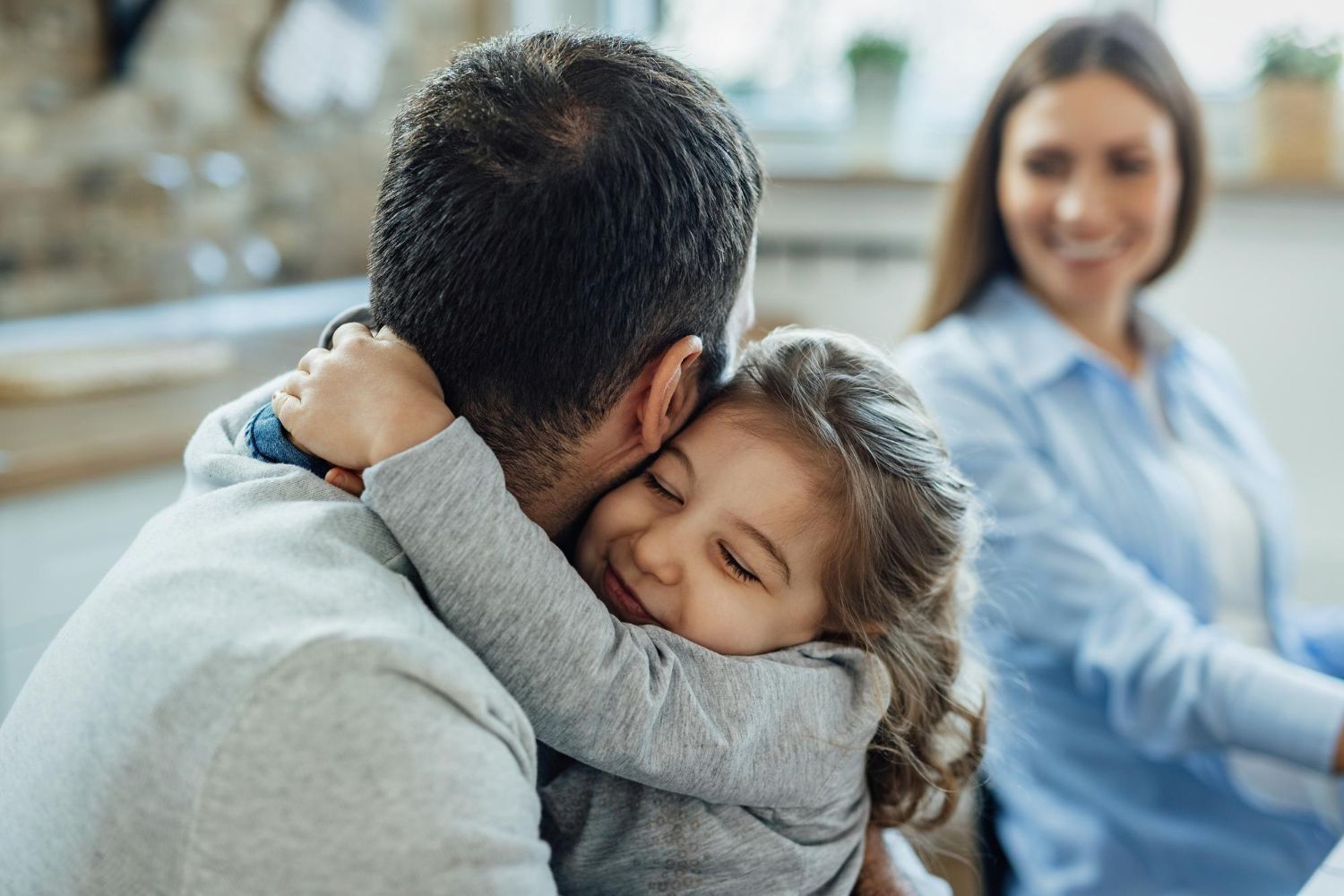 Child hugging an adult indoors, with a smiling woman blurred in the background