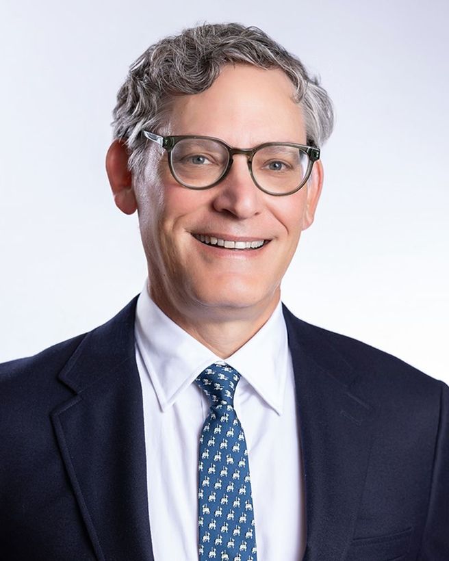 Portrait of a man in a navy suit, white shirt, and blue patterned tie, smiling against a light background