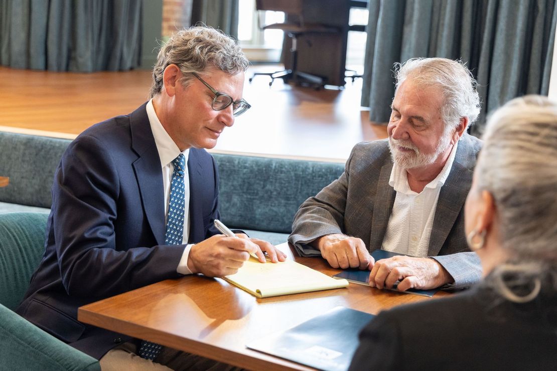 Two men in suits discuss documents at a conference table, with another person seated nearby.