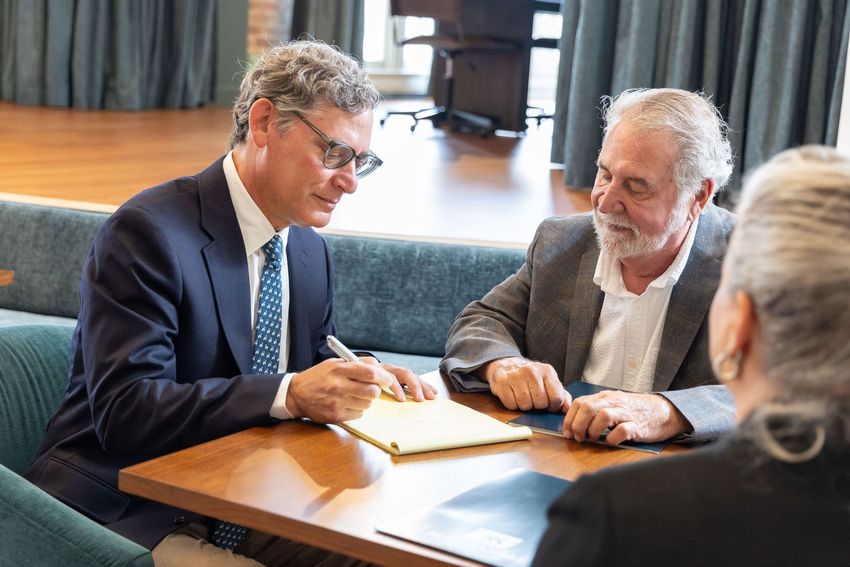 Two men in suits discuss documents at a conference table, with another person seated nearby.