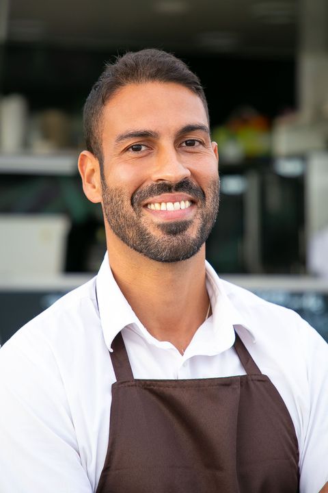 Smiling person in a white shirt and brown apron standing in front of a shop counter