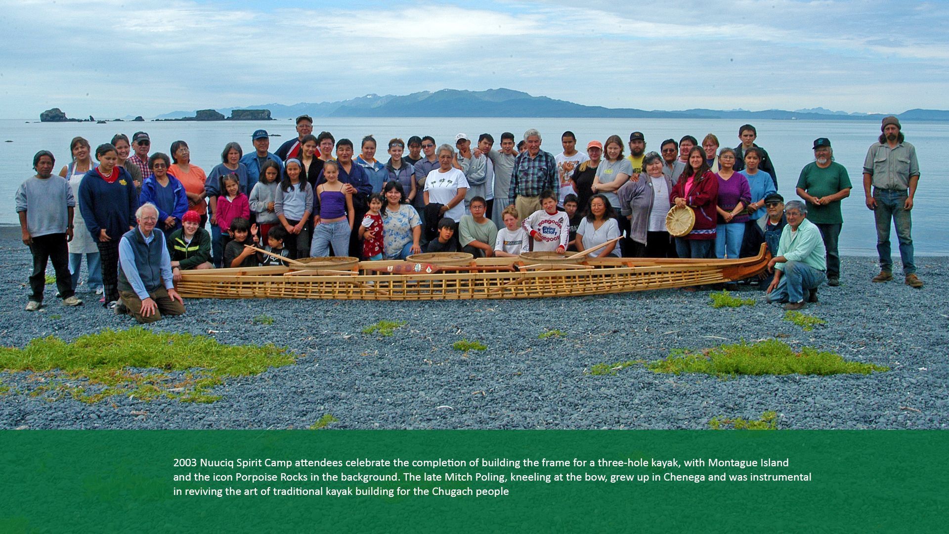 Group of people with a wooden boat on a beach, mountains in the background. Cloudy sky.