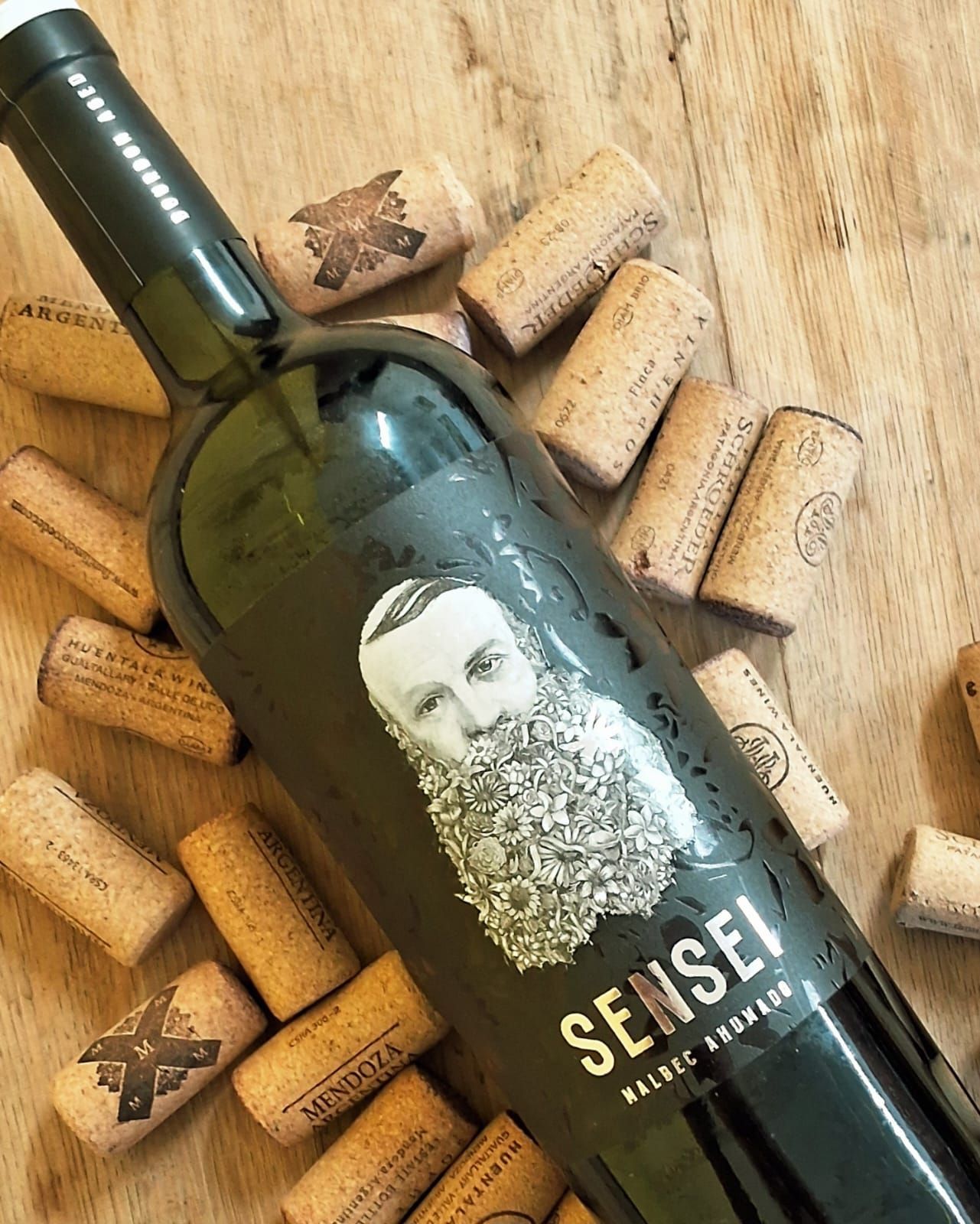 Una botella de vino Sensei Malbec colocada en diagonal sobre una superficie de madera, rodeada de corchos dispersos.