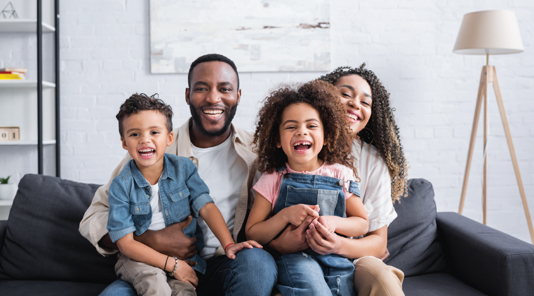 Family of four smiling on a couch in a living room; bright and happy.
