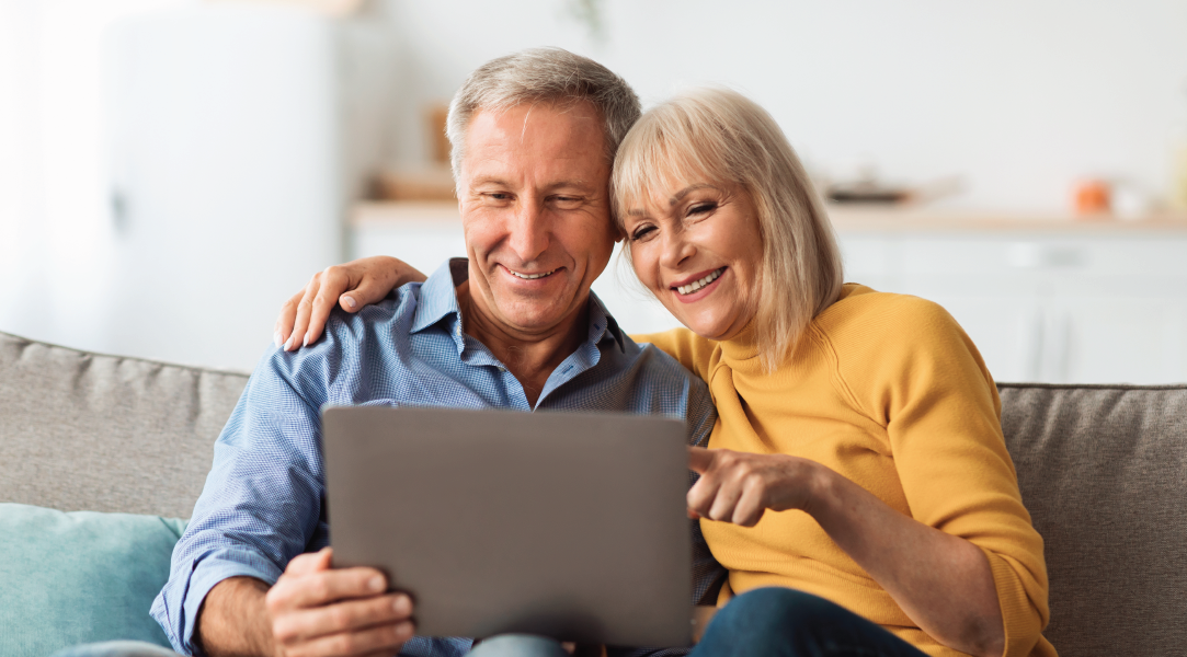 Older couple on couch looking at laptop, smiling. Woman points at screen; man has arm around her.