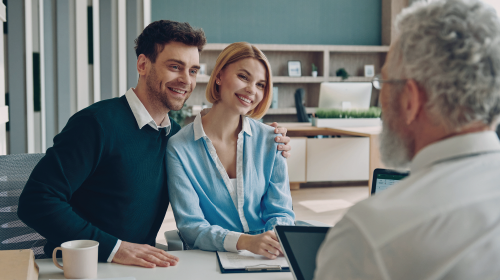Couple smiling, embracing at a table with a person; signing a document. Office setting, bright.