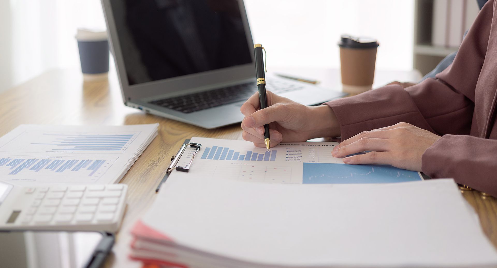 Person writing on documents at a desk with a laptop, calculator, and coffee cups.