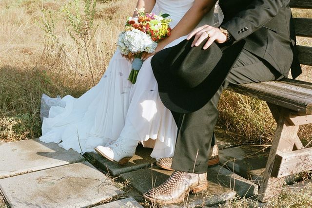 A bride and groom wearing cowboy boots sit on a wooden bench outdoors, holding a bouquet of colorful flowers.