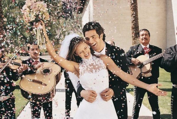 Newly married couple dances with joy as confetti falls, mariachi band plays outdoors.