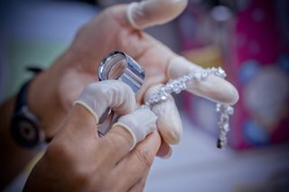 Hands in white gloves examine a diamond bracelet with a magnifying glass.