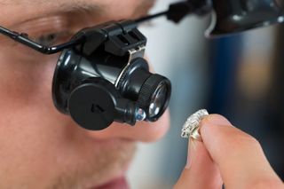 Person wearing magnifying glasses examining a small, possibly jeweled, object held in their fingers.