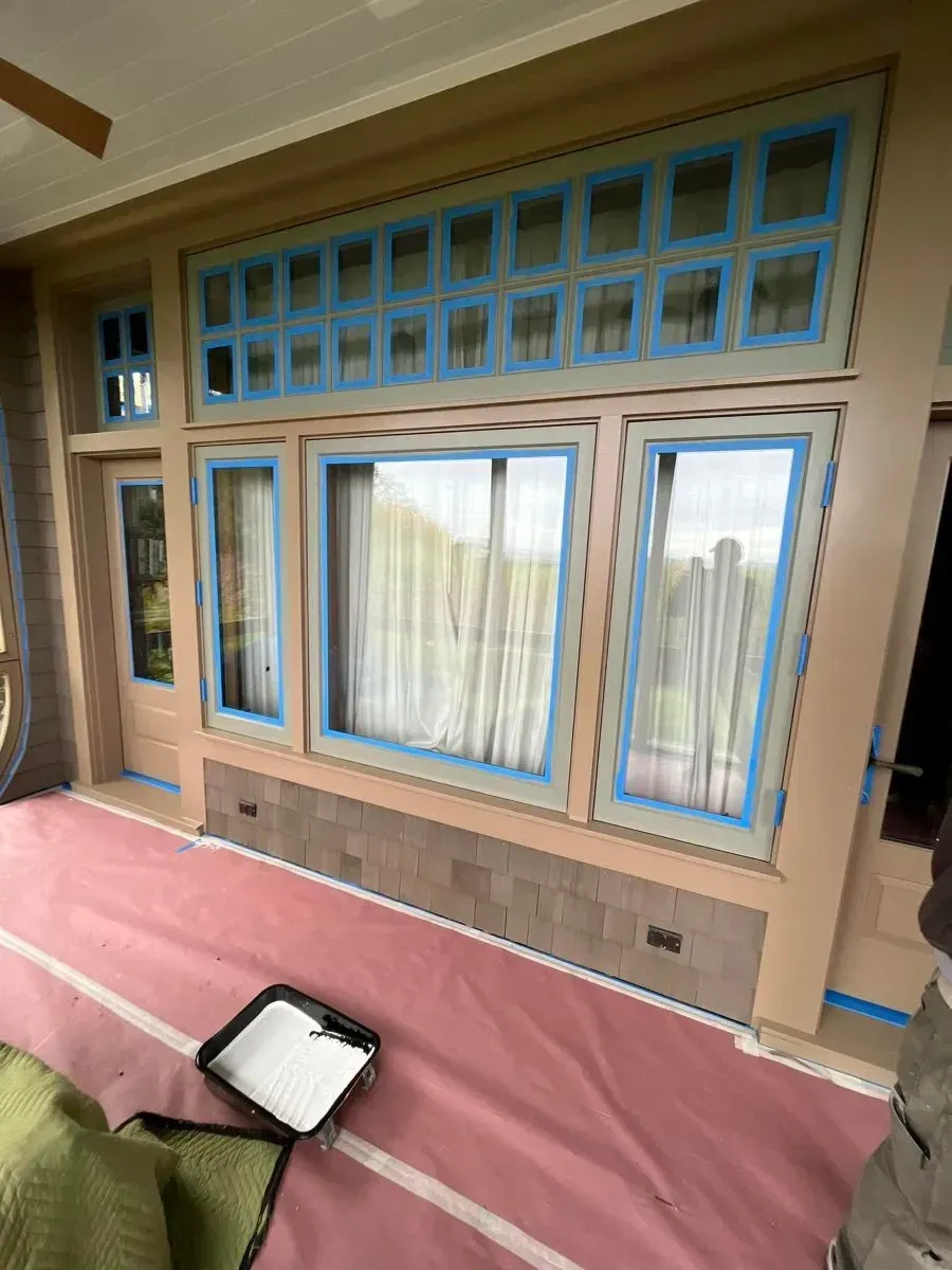 Covered porch with large blue-trimmed windows and sheer curtains above a pink floor covering.