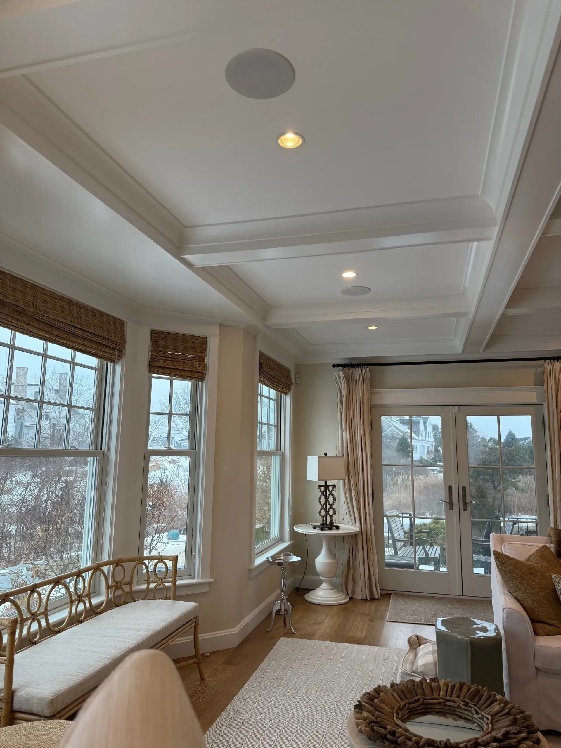 Bright sunroom with white coffered ceiling, wicker window seat, and large windows overlooking water.