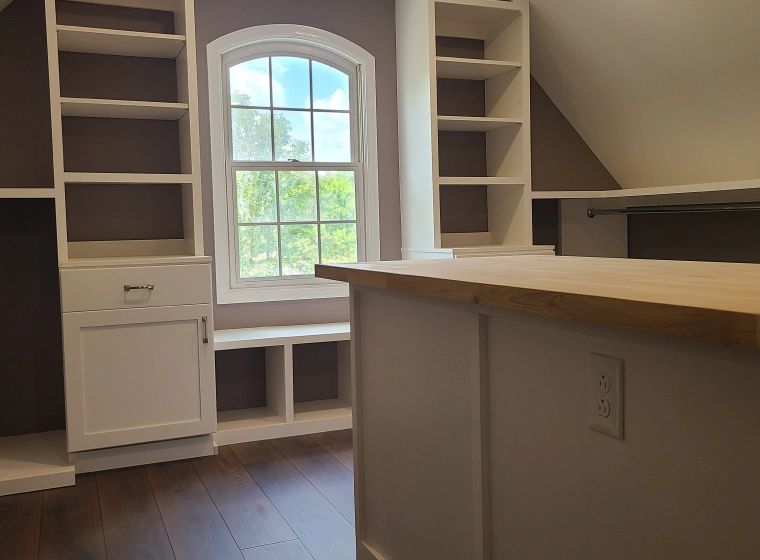 A custom white walk-in closet with built-in shelving, drawers, a window seat, and a central island with a wood countertop.