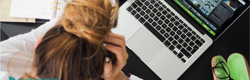 Person working at a laptop while holding their head, with green shoes visible on the floor.
