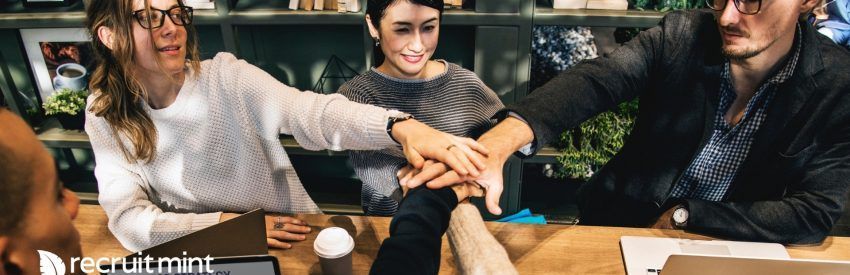 Three coworkers stacking hands in a cozy office lounge, smiling with laptops and coffee cups nearby
