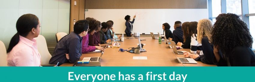 People seated around a conference table in a meeting room with a teal banner reading “Everyone has a first day”