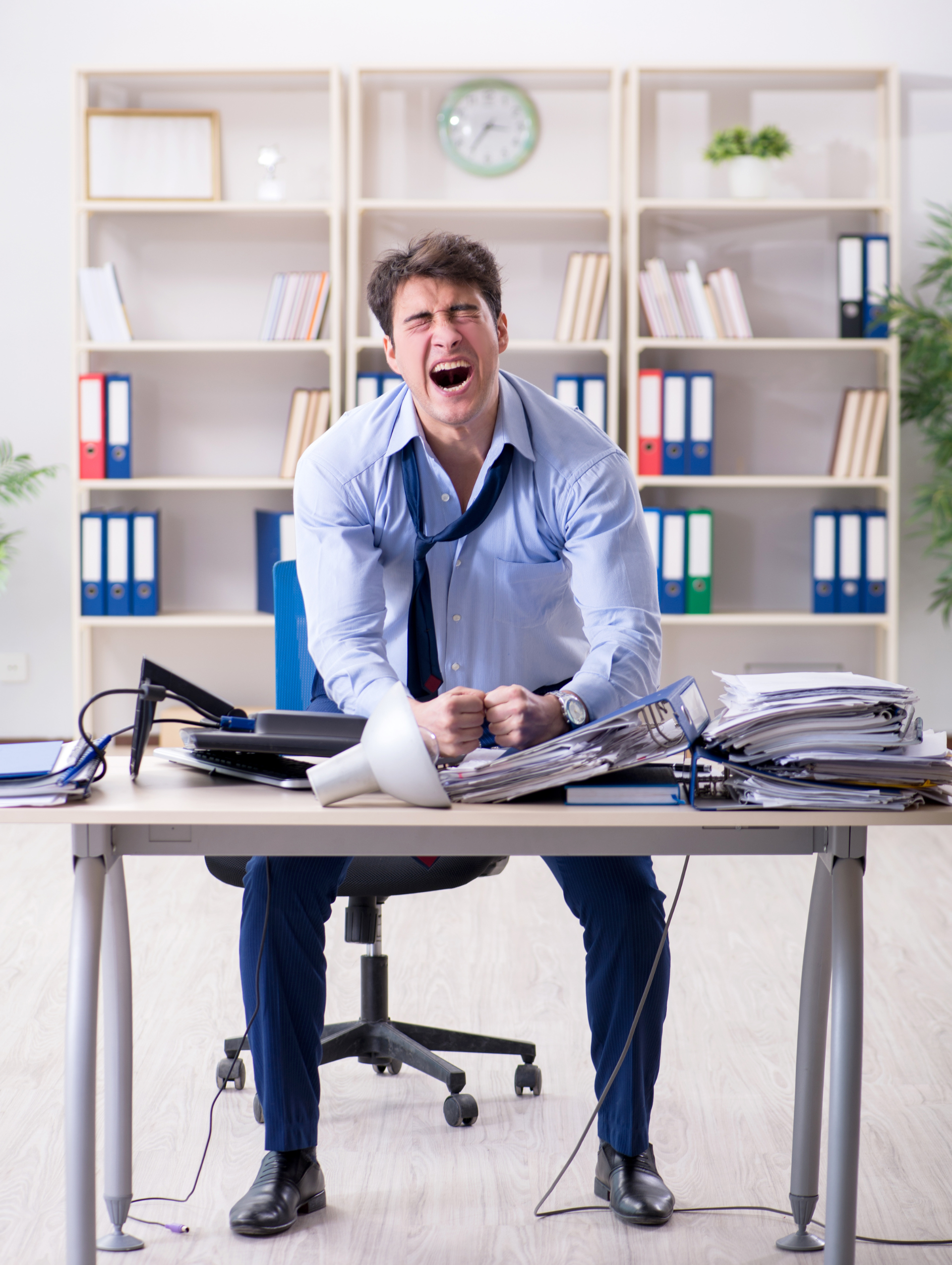 Frustrated man in blue shirt at office desk, with papers and laptop, shouting in a bright office