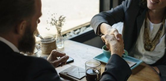 Two people in business attire shaking hands across a desk in a meeting room