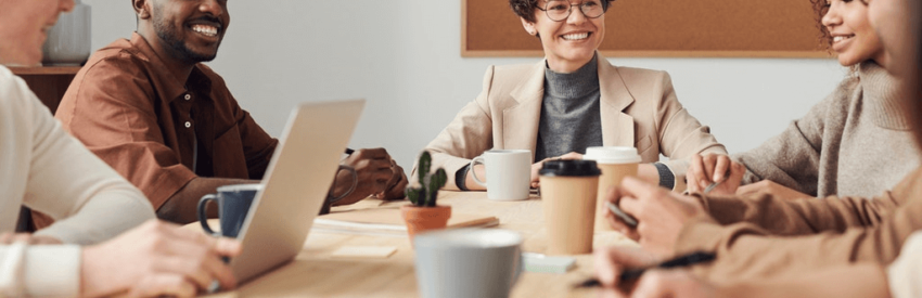 People gathered around a table in a bright office, smiling and talking with laptops and coffee cups.