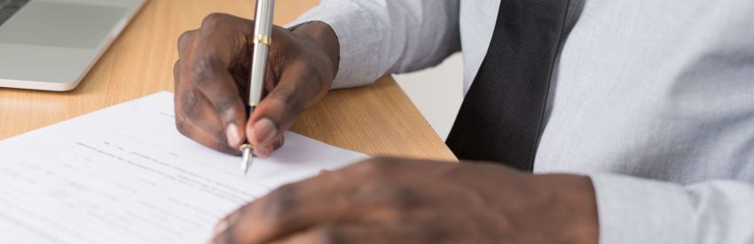 Person writing with a pen at a desk with papers and a laptop nearby