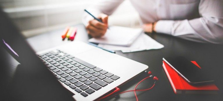 Laptop on desk with a person writing notes, red glasses, and a red notebook nearby