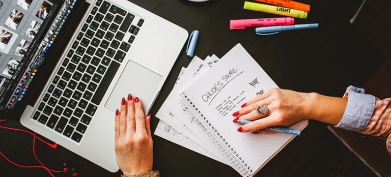 Hands typing on a laptop beside a notebook with notes and colorful pens on a desk