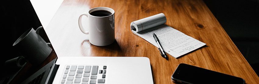 Laptop, coffee mug, notebook, and pen on a wooden desk with warm light.