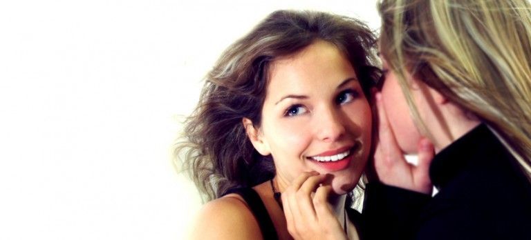 Two women smiling and whispering closely against a white background