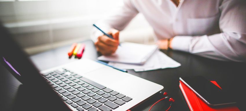 Person writing at a desk beside a laptop and red notebook