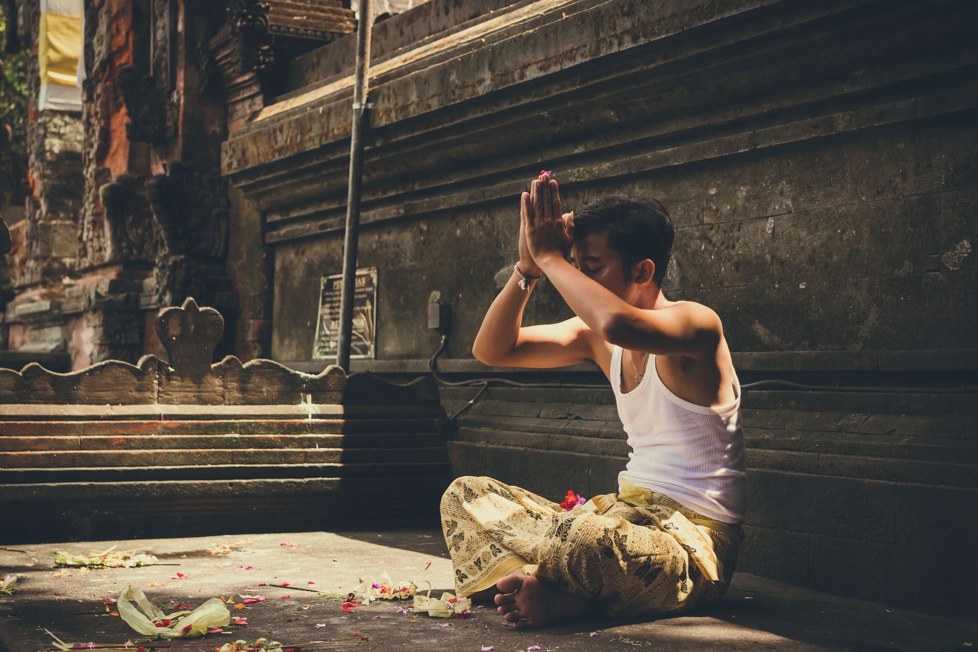 Man in white tank top and patterned pants, in prayer position, outside a temple. Why practice the 5 points of yoga.