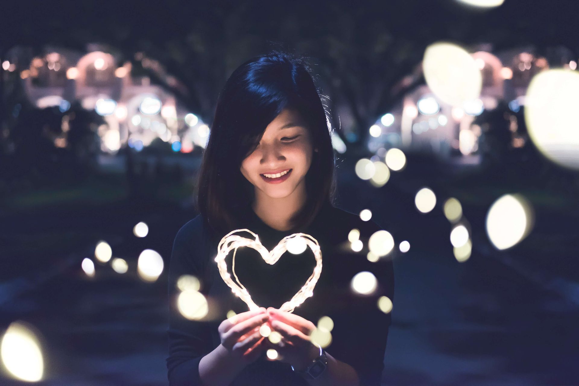 Woman holding glowing heart, smiling in a dark setting with bokeh lights.