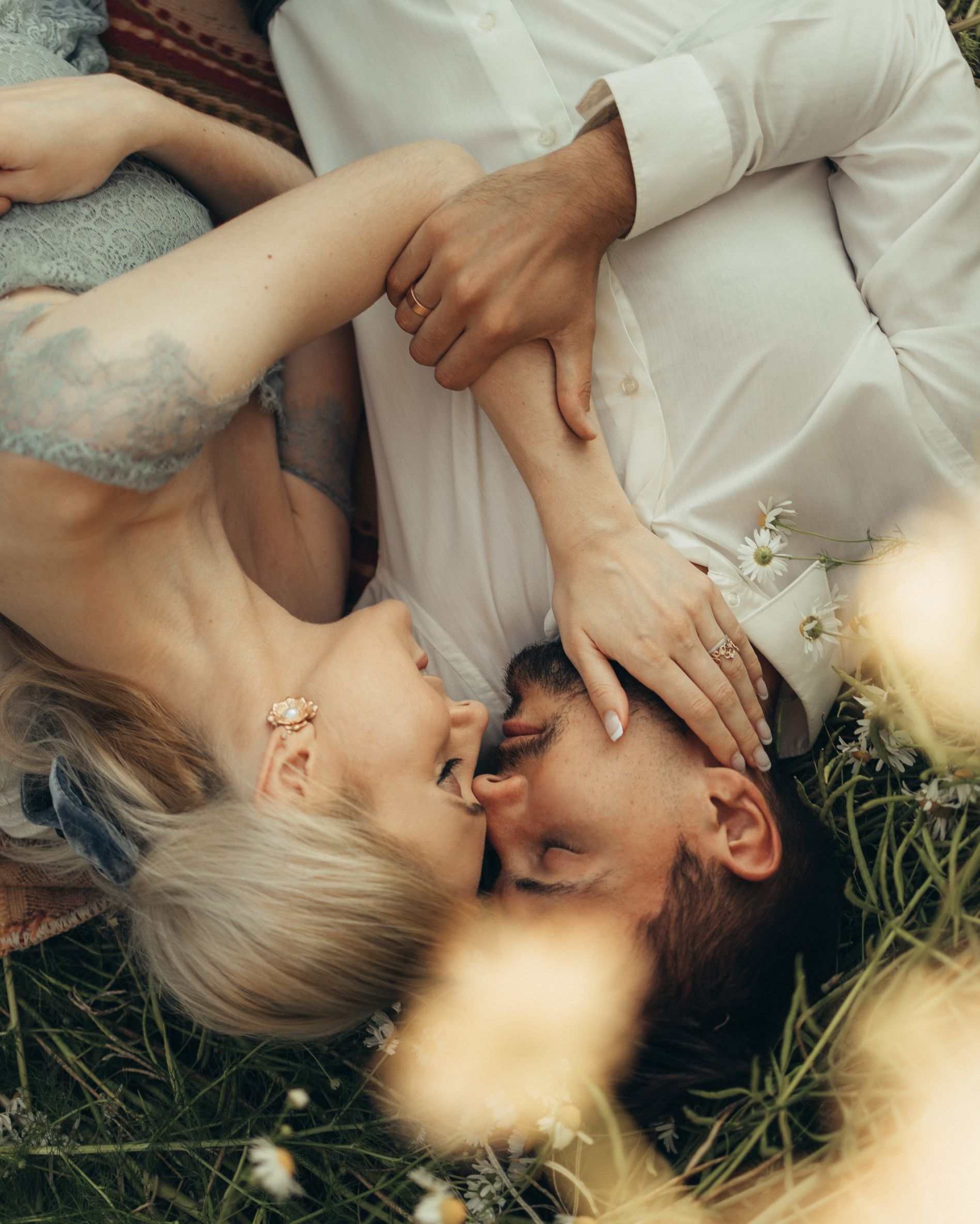 Couple in embrace, lying in grass; woman’s arm around man's neck. White and blue clothing, sunlight.