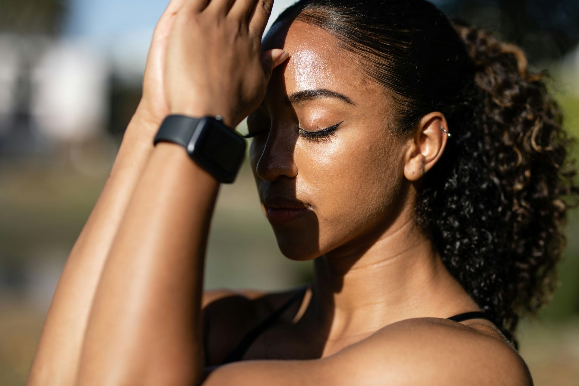 Woman with eyes closed, hands to forehead, wearing a fitness tracker, outside in the sun.