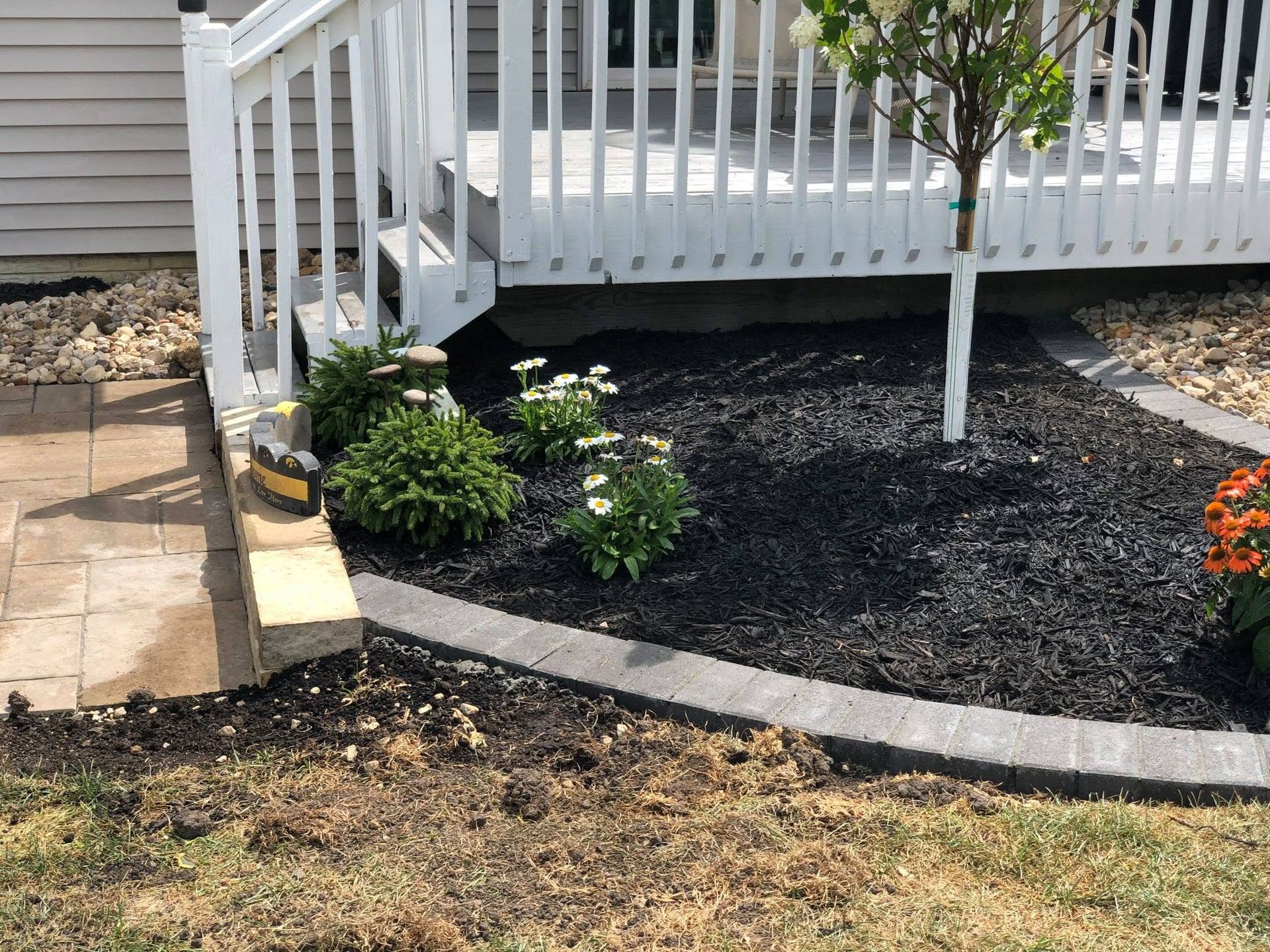 Small landscaped front yard with black mulch, shrubs, and a white porch railing beside stone pavers.