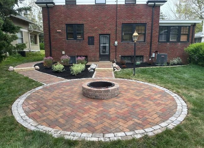 Backyard brick patio with a circular fire pit and white paver border in front of a brick house